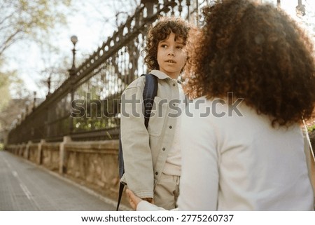 The son stands and looks away while his mother, who is crouched opposite, holds his hand