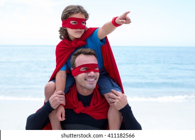 Son in superhero costume pointing while father carrying him on shoulder at beach - Powered by Shutterstock