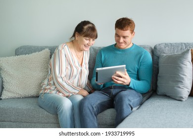 Son Teaching His Mother To Use Tablet. Older People Using Technology. Cheerful Elderly Woman Sitting On The Sofa Next To His Adult Son