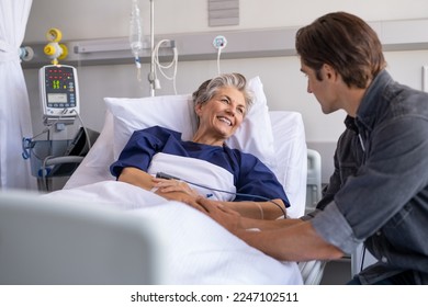 Son visiting sick old mother in hospital. Smiling senior patient in conversation with man. Grandson visiting and cheering his happy granny lying in bed at modern hospital ward. - Shutterstock ID 2247102511