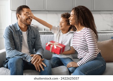 Son And Wife Greeting Young Black Man With Father's Day, Giving Gift At Home. Male Kid Covering Dad's Eyes, Surprising With Present, African American Family Sitting On Couch In Living Room, Closeup - Powered by Shutterstock