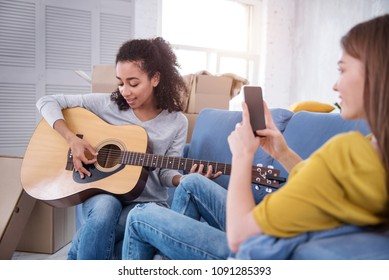 Song For You. Upbeat Curly-haired Girl Playing Guitar And Dedicating A Song To Her Roommate While She Filming Her On Phone