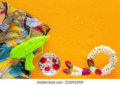 Songkran festival background with colorful shirt, water gun, flowers in water bowl and jasmine garland put on clear mirror that have wet yellow background below.