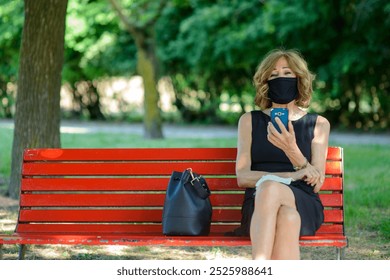 Sophisticated businesswoman taking a break in city park. Checking smartphone with face mask. Blending work and relaxation. Professional style in nature. Emphasizing health and safety during pandemic - Powered by Shutterstock