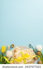 Sophisticated Passover theme: top-view vertical photo showcasing matzah wrapped in ribbon, nuts, egg, honey dipper, nutcracker, and tulips on a gentle blue background, space for message