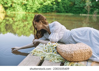 a sophisticated woman in a light dress touches the water while relaxing by the lake