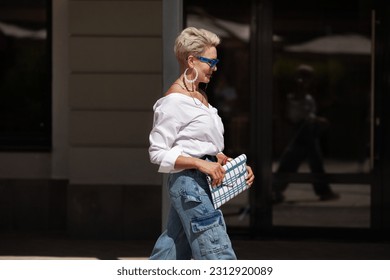 Sophisticated woman with short hair in white shirt and cargo jeans pants, fashionable accessories earrings, stylish glasses and clutch walking outdoors in city. Close up Fashion style.