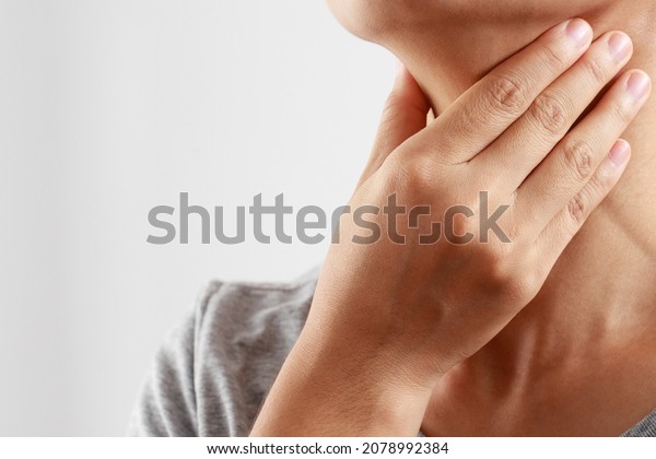 sore throat pain. Closeup of young woman sick\
holding her inflamed throat using hands to touch the ill neck in\
blue shirt on gray background. Medical and healthcare concept.\
Focus red on to show pain.