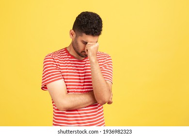 Sorrowful hopeless man in striped t-shirt lowering his head hiding eyes, upset depressed about mistake, become broke, feeling shame. Indoor studio shot isolated on yellow background