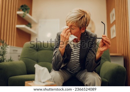 A sorrowful senior woman is crying and wiping her tears while sitting in the doctor's office during her session with a shrink. A sad and depressed senior woman is expressing pain and sorrow.