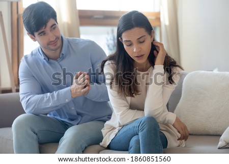 I am so sorry. Guilty millennial man husband partner of young woman sitting on sofa behind her back with clasped hands asking forgiveness. Male excusing wish to reconcile after quarrel conflict