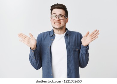 Sorry I messed up. Guilty young and handsome male in glasses and blue shirt raising hands near shoulders, shrugging making awkard smile as apologizing for making mistake over white background