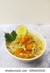 Soto ayam is served complete with shredded chicken, vegetables, crispy potatoes, and a sprinkling of fried onions. Served in a bowl with lime wedges
