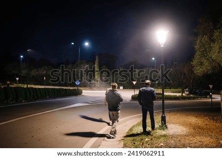 Sotogrande, Spain - January, 23, 2024 - Three people stand by a lamppost on a well-lit road at night with trees and a car passing by.