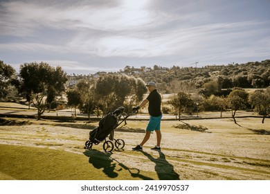 Sotogrante, Spain - January 25, 2024 - A golfer pushing a golf bag trolley on a course with trees and a hilly landscape in the background. - Powered by Shutterstock