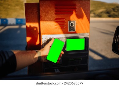 Sotogrante, Spain - January 26, 2024 -  hand reaching out from a car window holding a smartphone with a green screen towards a toll booth machine. - Powered by Shutterstock
