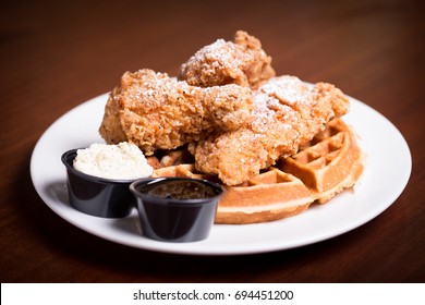 Soul food close up of fried chicken drumsticks with crispy and flaky breaded skin on fluffy Belgian waffle with side container of maple syrup and whipped butter dusted in powered confectioner sugar  