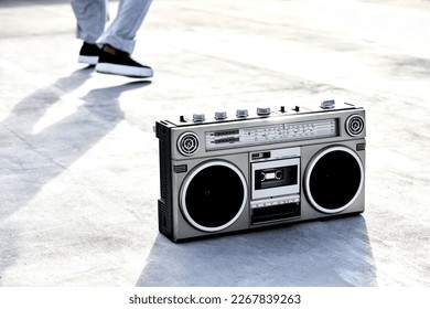 The sound of music. High angle shot of a boombox on the floor with an unrecognizable female dancer in the background.