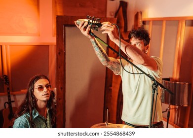 Sound technician is standing at recording music studio and adjusting microphone for drums recording while drummer is sitting and looking at the camera. Professional musician is preparing for recording - Powered by Shutterstock