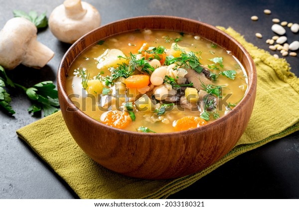 Soup with barley,\
beans, mushrooms, potatoes, carrots, celery, and herbs. Barley soup\
with vegetables in a wooden bowl on a black background. Vegan food,\
homemade soup.