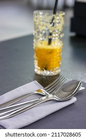Soup spoon and metal fork lie on a gray napkin on a table top view. A glass of fresh orange juice in morning. Table setting, cloth in restaurant, cafe, kitchen. Gray textile towel. Flatware close up.