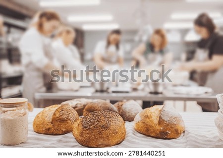 Sourdough bread sits on a table in the kitchen as bakers focus on their tasks. They knead dough and learn methods to create artisan bread. The scene shows a busy kitchen environment.