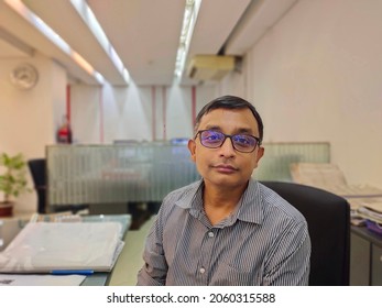 A South Asian Middle Aged Man Sitting On A Chair At Office,   Blurred Background.