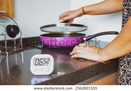 A South Asian woman covering the lid of frying pan on induction hob. Digital kitchen timer on modern kitchen table for multitasking. Selective focus on the timer.