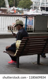 South Banks, London, UK Sept 2018. Man Looking At His Phone Wearing An Iconic Deer Stalker Hat, Sitting On A Bench, Earphones On, Isolated