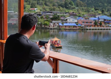 South East Asian Man Take Photo By Mobile Phone. Southeast Asian Adult Snap Picture For Village Scene With Mountain, Lagoon And Boat Tour. Male Sitting Close To Window At Northern Of Thailand.