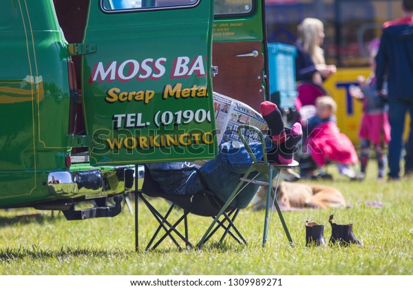 South Shields / Great Britain\
- June 5, 2016: Man sits on camping chairs reading a newspaper. \
Boots on ground, feet and socks on a stool.  Hiden behind vehicle\
doors.