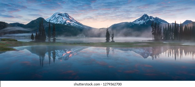 South Sister and Broken Top reflect over the calm waters of Sparks Lake at sunrise  in the Cascades Range in Central Oregon, USA in an early morning light. Morning mist rises from lake into trees.