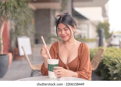 A Southeast Asian Lady Sitting Outside The Coffee Shop Putting A Paper Straw Into Her Coffee.