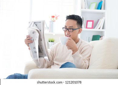 Southeast Asian Male Drinking Coffee While Reading News Paper Sitting On Sofa At Home, Indoor Lifestyle