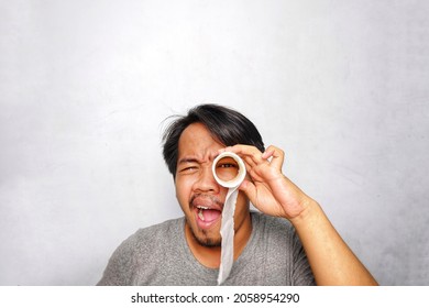 A Southeast Asian Man Peeping His Eye Through Hole Of Toilet Paper Roll And Wearing Grey Shirt