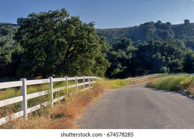 Southern California Rural Landscape, With Country Road Lined With Fence, Oak Trees And Mountains Background.