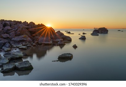 Southern coast of Finnish Gulf. Granite rocks piled. Sunrise time,  star beams behind boulders. Smooth transparent water, dark rocks. Blue hour turning to golden period. Estonia, Baltic