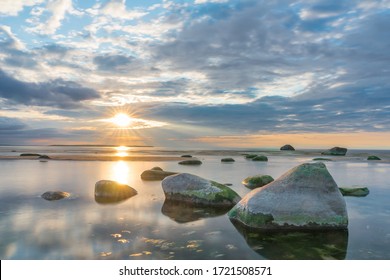Southern coast of the Finnish Gulf. Rocks covered with green seaweed in the Baltic sea. Smooth transparent reflective water. Orange sunset markings under the low altitude clouds. Estonia, Baltic
