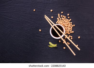 Soy sauce in a bowl with bamboo chopsticks and soya beans scattered on the black surface of the slate stone, top view, closeup with space for text - Powered by Shutterstock