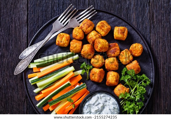 soya bean and chickpea mini rolls or gnocchi served
with fresh cucumber and carrot sticks and yogurt based sauce with
herbs on black plate on dark oak wood table, horizontal view from
above, flat lay