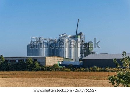 A soybean processing facility is being constructed, featuring tall silos and industrial buildings. The site is surrounded by open fields, with trees lining the outskirts.