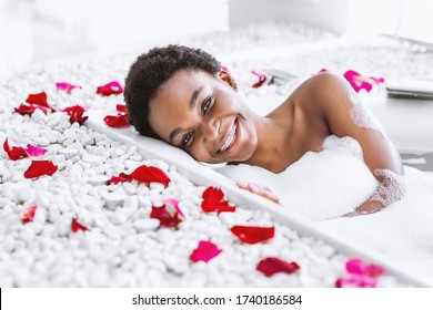 Spa day and beauty care at home. Smiling african american girl takes bath with foam and petals