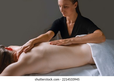Spa procedure in a massage room, a woman patient lies relaxed on the table covered with a white terry towel, gentle hands of a female massage therapist massage her back