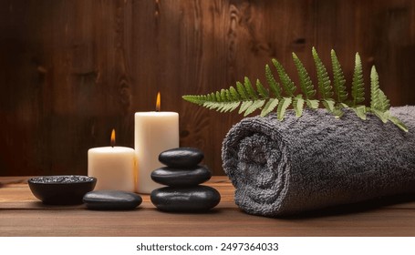 A spa setting featuring a gray towel rolled up beside lit candles and smooth stones. Fern leaves and a small bowl of black pebbles add to the serene atmosphere, set against a warm wooden background. - Powered by Shutterstock