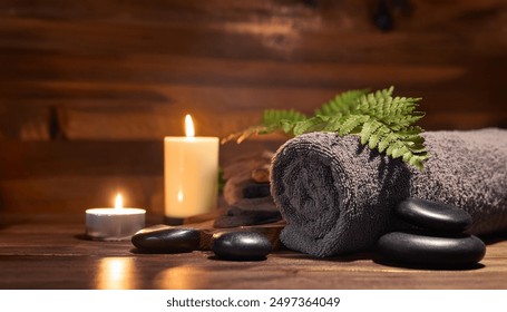 A spa setting featuring a gray towel rolled up beside lit candles and smooth stones. Fern leaves and a small bowl of black pebbles add to the serene atmosphere, set against a warm wooden background. - Powered by Shutterstock