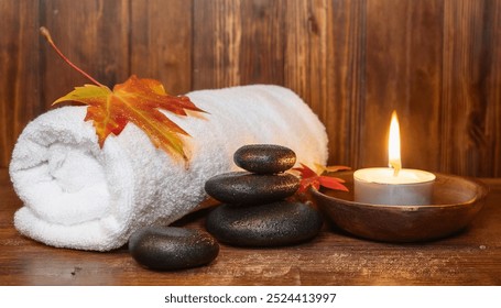 A spa setting featuring a white towel rolled up beside lit candle and smooth black stones. Autumn leaves and a small bowl of black pebbles add to the serene atmosphere. - Powered by Shutterstock