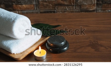 Spa setup featuring stacked rolled white towels on a wooden tray, stacked dark smooth massage stones, a glowing burning candle, and green leaves on a dark wooden surface with a stone wall background.