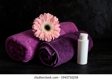 Spa Still Life. Two Purple Towels, A Bottle Of Cream And A Pink Gerbera On The Table Close-up