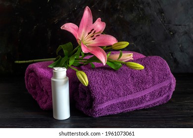 Spa Still Life. Two Purple Towels, A Bottle Of Cream And A Pink Lily On The Table Close-up