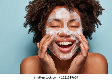 Spa, Therapy And Beauty Concept. Satisfied Black Woman Washes Face With White Soap, Has Cleansing Foam, Purification Of Face, Cleans Pores, Shows Bare Shoulders, Isolated Over Blue Studio Wall